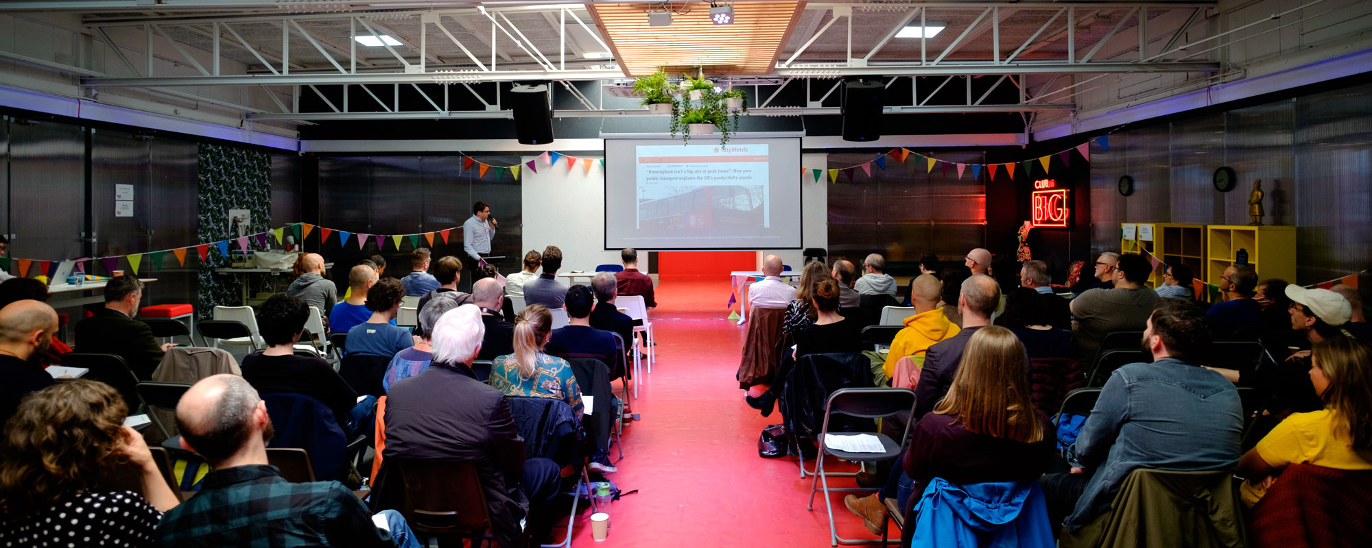 View from the audience at the Festival of Maintenance, taking place a low, wide event space with a red floor and a projection screen in the middle