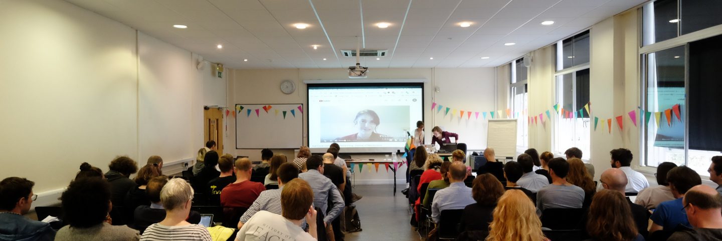 View from the audience at the Festival of Maintenance, taking place in a university classroom, with multi-coloured flag bunting hung up across the walls